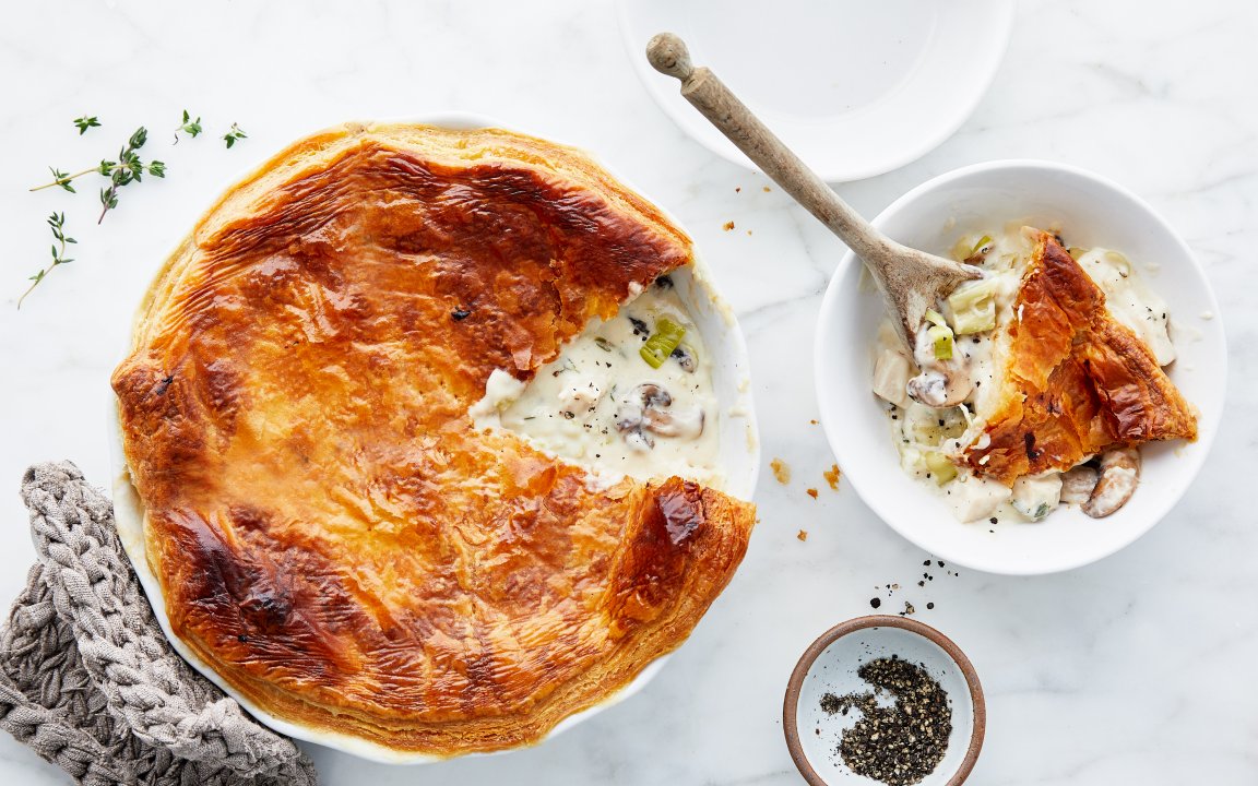 Golden, flaky pot pie with a rich and creamy filling of chicken, mushrooms, and leeks, served in a baking dish with a portion scooped into a bowl. A wooden spoon rests in the bowl, and a small dish of cracked black pepper adds a finishing touch. Fresh thyme sprigs and a cozy potholder complete the rustic presentation on a light marble table.