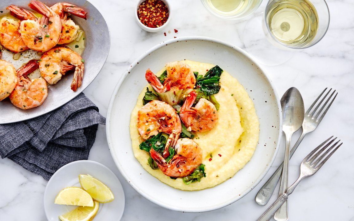 Shrimp and grits plated beautifully with creamy polenta, sautéed greens, and perfectly seared shrimp, garnished with a sprinkle of red pepper flakes. A plate of additional shrimp, lemon wedges, and a small bowl of chili flakes are nearby, accompanied by two glasses of white wine on a light marble surface for a sophisticated dining experience.