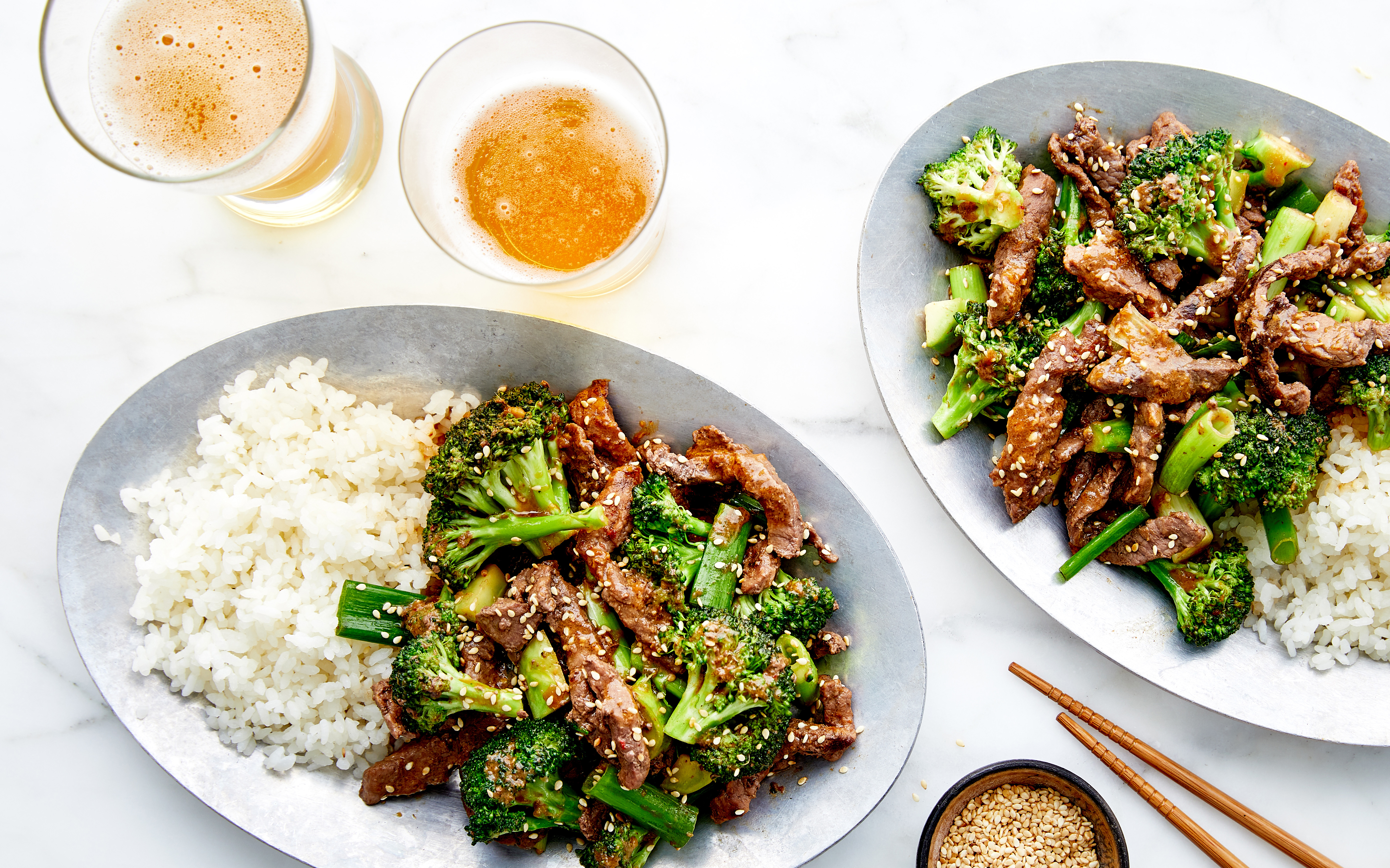 Two plates of beef and broccoli stir-fry served with steamed white rice, topped with sesame seeds for added flavor. The vibrant dish is complemented by two glasses of beer and a small bowl of sesame seeds on the side. Wooden chopsticks complete the setting on a light marble table, offering an inviting, balanced meal.