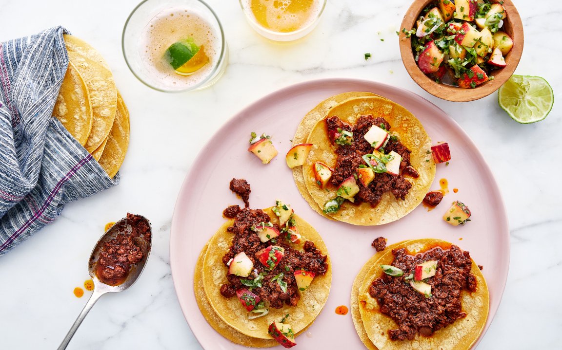 Three corn tortillas topped with a savory chorizo mixture and a colorful peach salsa, served on a pink plate with lime wedges and a glass of beer.
