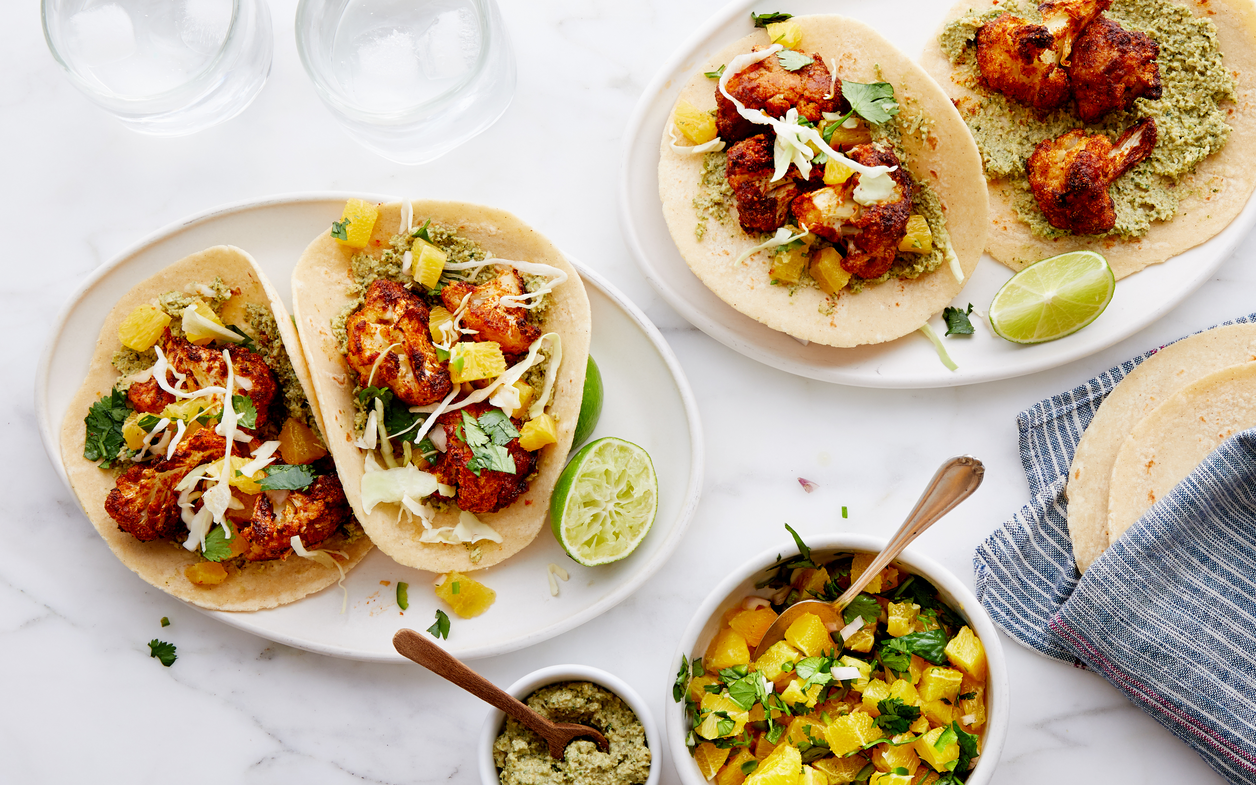 Colorful fish tacos served on soft tortillas, topped with crispy spiced fish, fresh cabbage, citrus salsa, and cilantro, paired with a creamy green sauce. Lime wedges, extra tortillas, and bowls of salsa and sauce accompany the meal, set on a light marble surface for a vibrant, fresh presentation