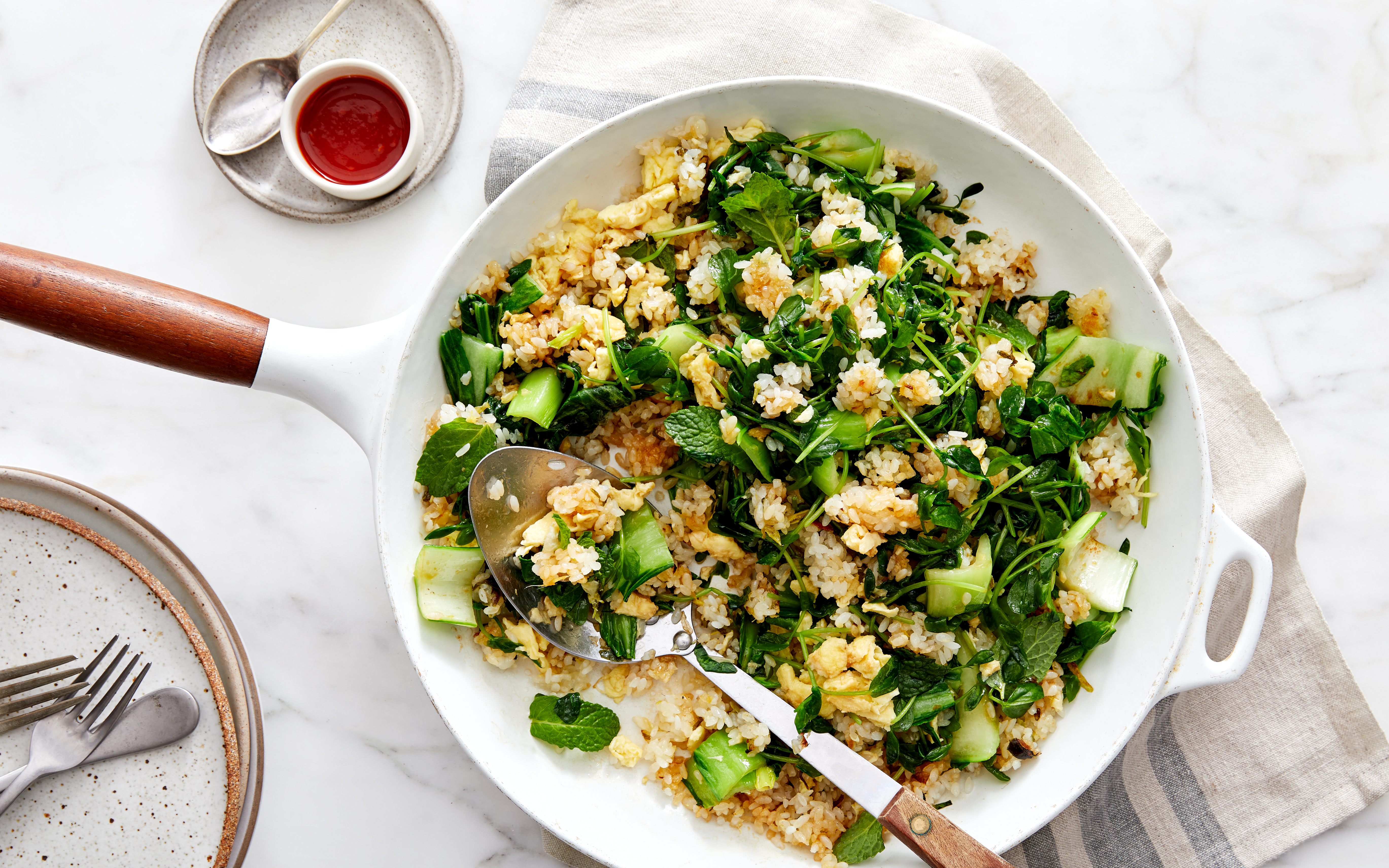 Stir-fried rice with bok choy, mint, and watercress in a white skillet, served with a side of Sriracha sauce.