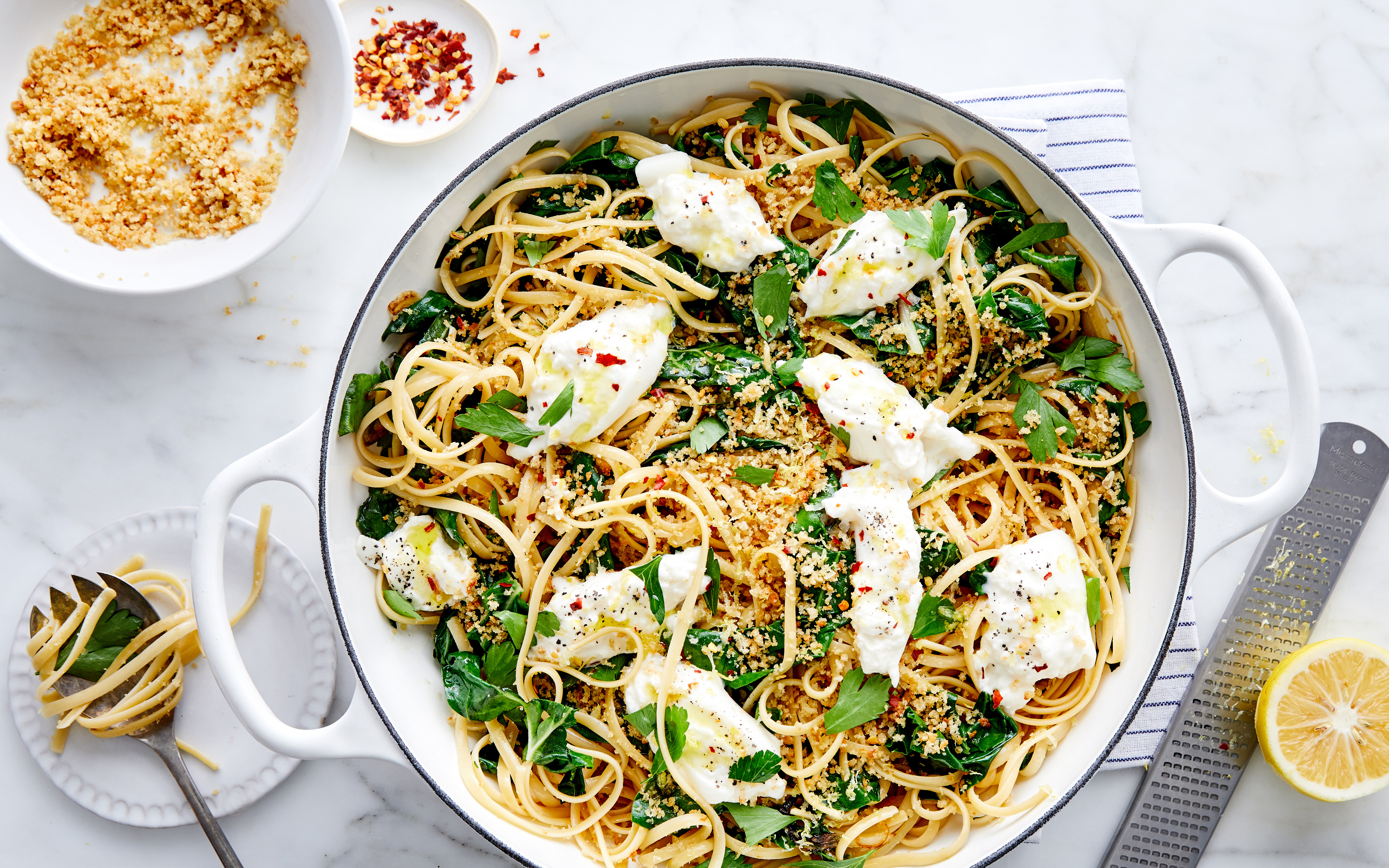 A large white skillet filled with linguine pasta, topped with dollops of fresh burrata, crispy breadcrumbs, parsley, and a sprinkle of chili flakes. A zested lemon, plate with a forkful of pasta, and extra breadcrumbs are placed around the skillet on a marble surface.