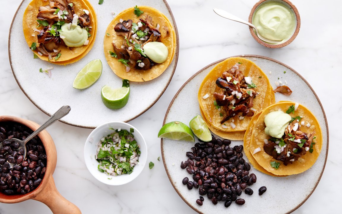 Three plates of mushroom tacos served on corn tortillas, garnished with fresh cilantro, diced onions, and a dollop of avocado crema. Accompanied by black beans, lime wedges, and a bowl of chopped herbs on a white marble surface. A wooden bowl with black beans and a spoon is visible in the foreground.