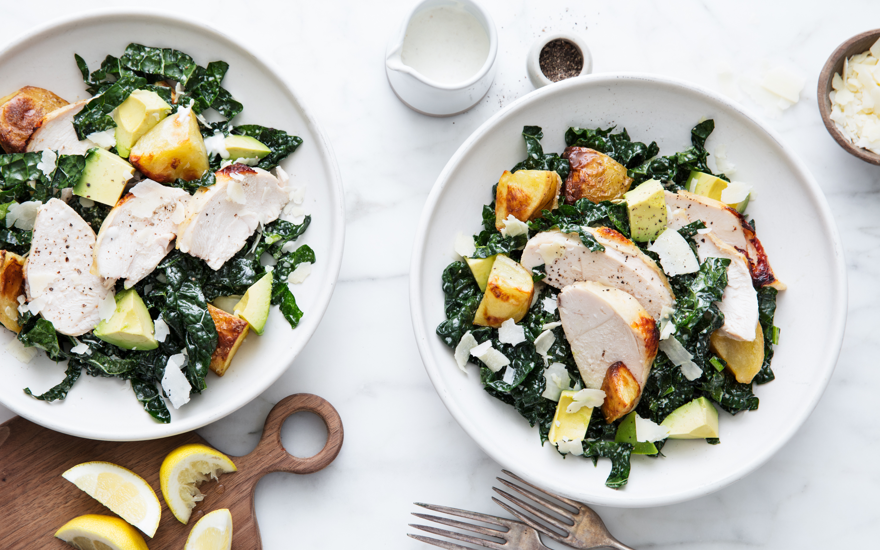 Grilled chicken and kale salad with roasted potatoes, avocado, shaved parmesan, and lemon wedges, served in white bowls on a marble countertop.