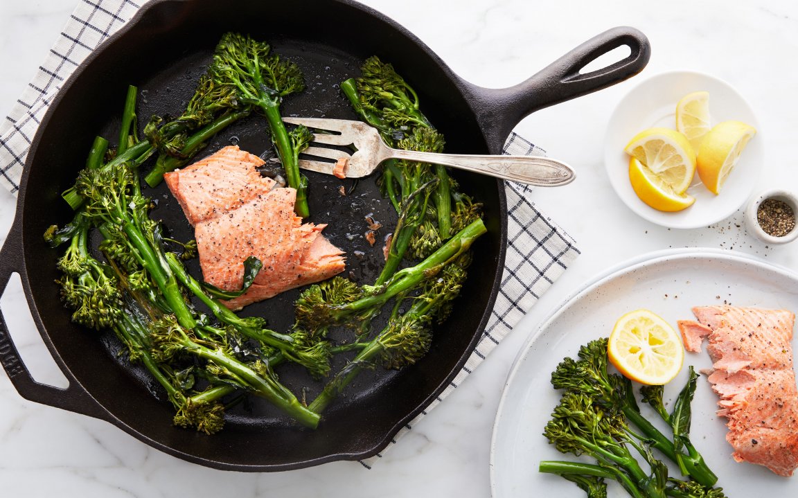 Pan-seared salmon with roasted broccolini in a cast iron skillet, served with fresh lemon wedges on a marble countertop.