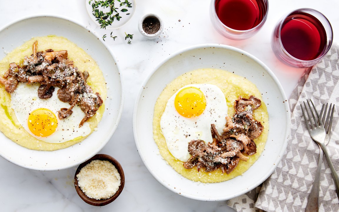 Two plates of creamy polenta topped with sunny-side-up eggs and sautéed mushrooms, sprinkled with grated Parmesan cheese. Fresh thyme, cracked pepper, and two glasses of red wine complete this elegant and comforting meal, served on a light marble surface with a patterned napkin and fork nearby.