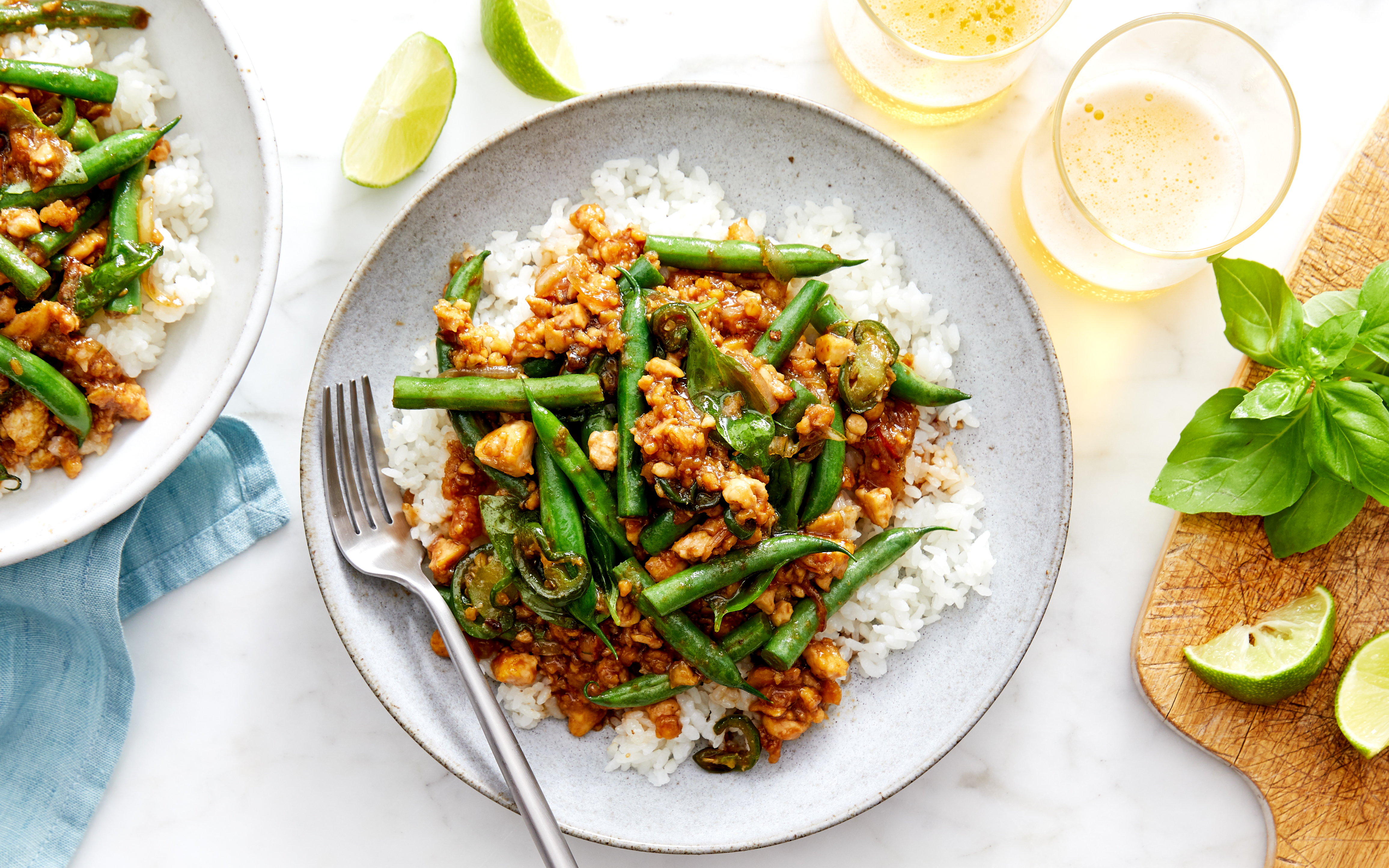 Spicy basil chicken stir-fry with green beans and jalapeños over white rice, served in a gray ceramic bowl with lime wedges and fresh basil on a wooden cutting board.