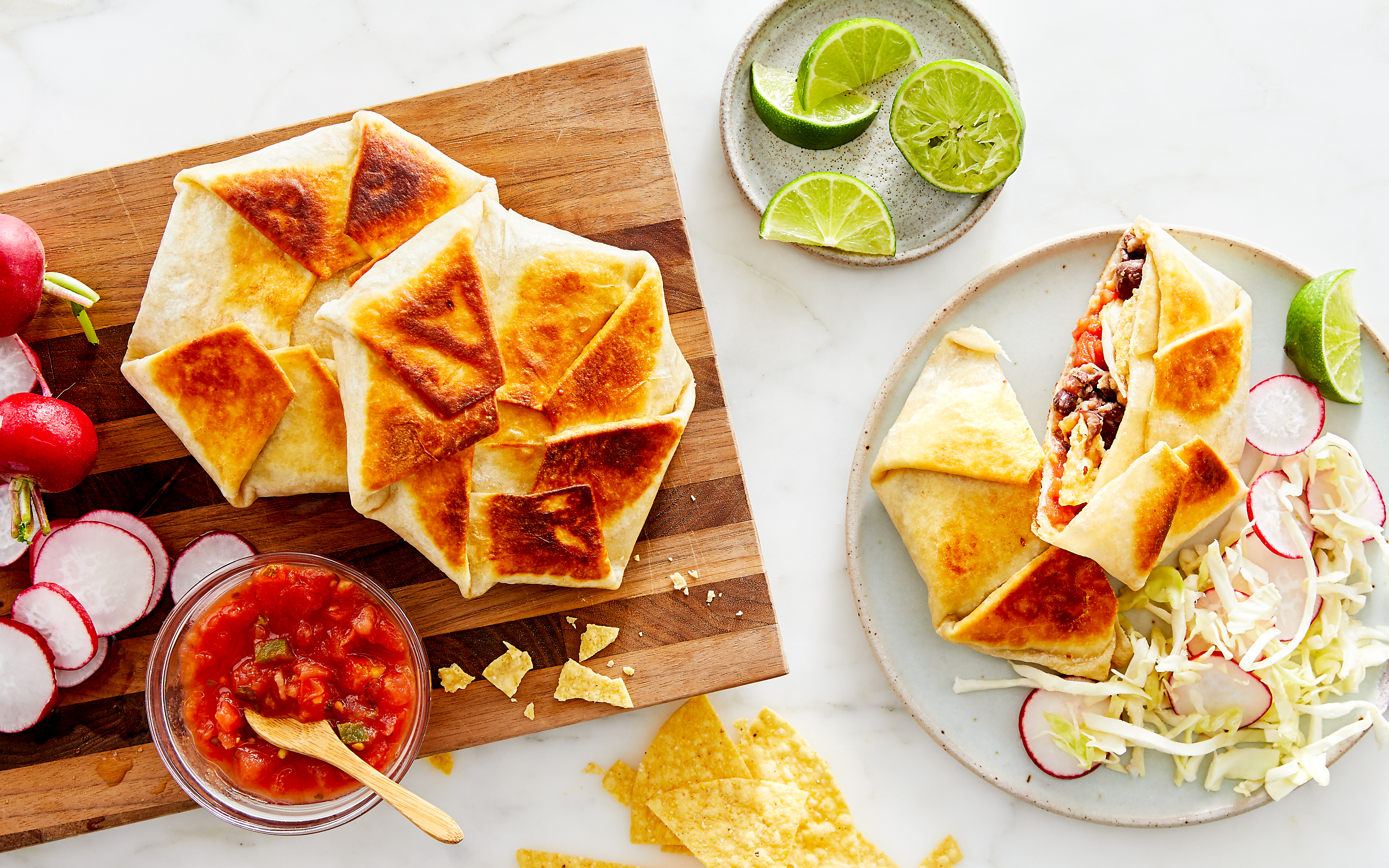 Crispy, golden-brown folded burritos on a wooden cutting board with radish slices, salsa in a glass bowl, and tortilla chips. A plated burrito, cut open to reveal beef, eggs, and vegetables, sits next to shredded cabbage, radish slices, and lime wedges on a marble surface.