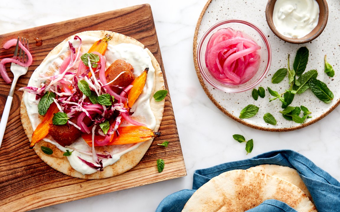 Grilled pita topped with roasted carrots, pickled red onions, shredded cabbage, fresh mint, and a creamy yogurt sauce, served with a side of extra toppings including pickled onions and fresh mint on a white plate.