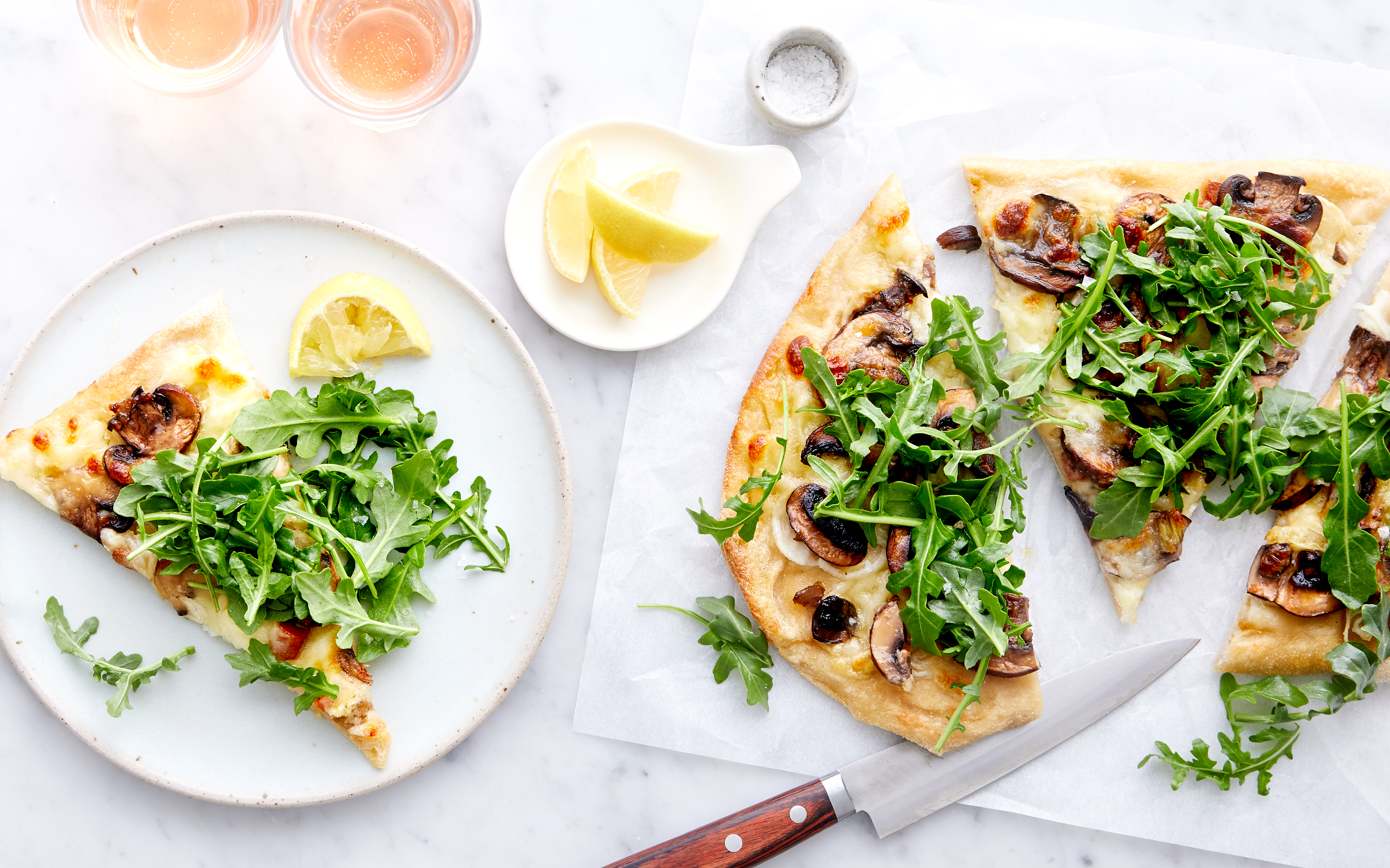 Flatbread pizza with mushrooms, caramelized onions, and fresh arugula, sliced into pieces on parchment paper. A plated slice with arugula sits beside a dish of lemon wedges. A sharp knife and glasses of rosé wine are also in the scene.