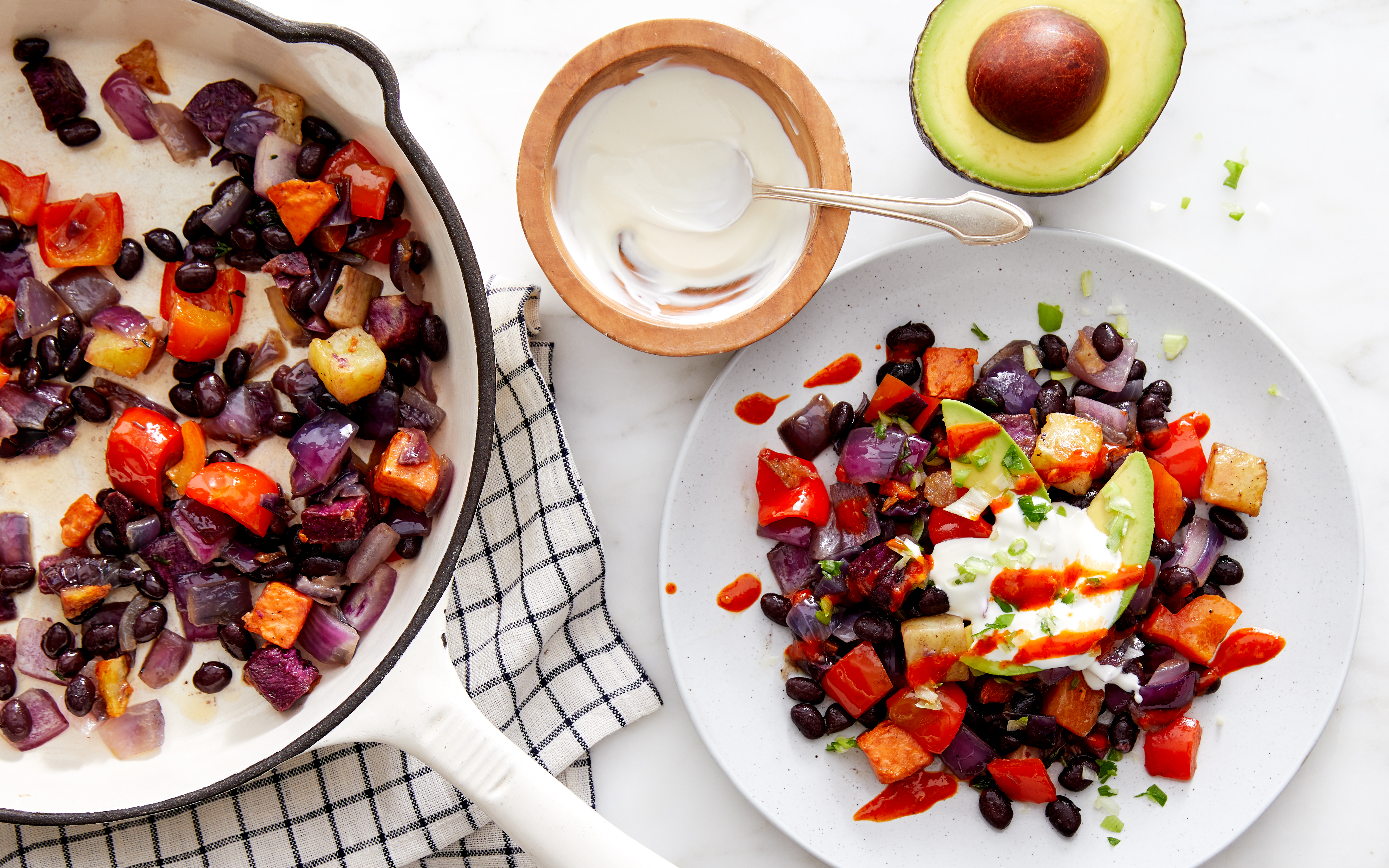 Vibrant vegetable hash featuring roasted sweet potatoes, purple potatoes, black beans, and red peppers, served with a dollop of sour cream, sliced avocado, and a drizzle of hot sauce. A skillet with the roasted vegetables, a bowl of sour cream, and a halved avocado complete the colorful and hearty meal setup on a light marble surface.