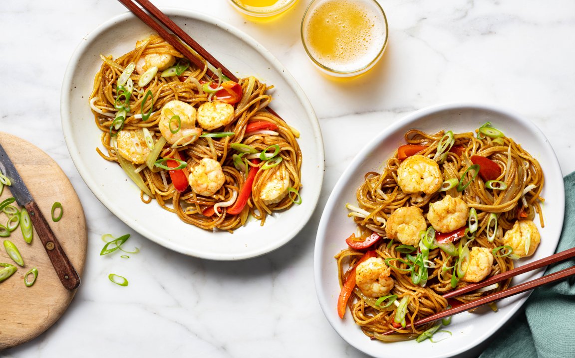 Stir-fried shrimp lo mein with bell peppers, scallions, and soy sauce, served in white oval plates with chopsticks on a marble countertop.